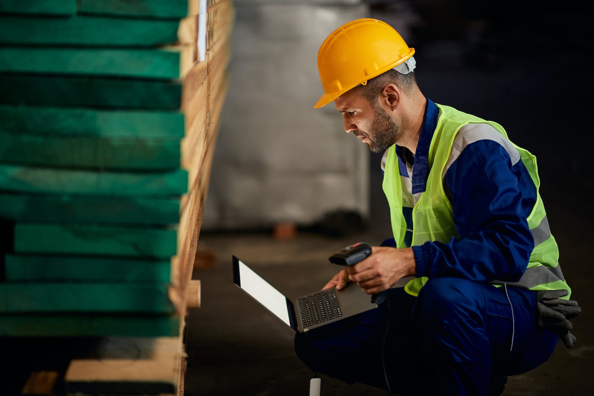 Warehouse worker using laptop and bar code scanner at lumber storage compartment.